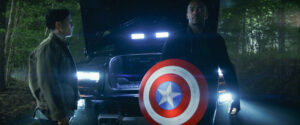 Two people stand in front of a truck with headlights on in a wooded area. One holds a round shield with a star in the center.