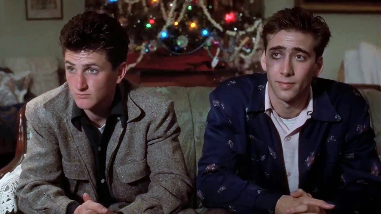 Two young men sit on a couch in front of a decorated Christmas tree, wearing casual jackets.