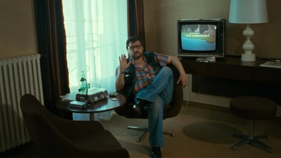 A person sits in a chair, waving, with a TV showing "Merry Christmas" in the background. There's a table with a bottle, two glasses, and a lamp nearby.