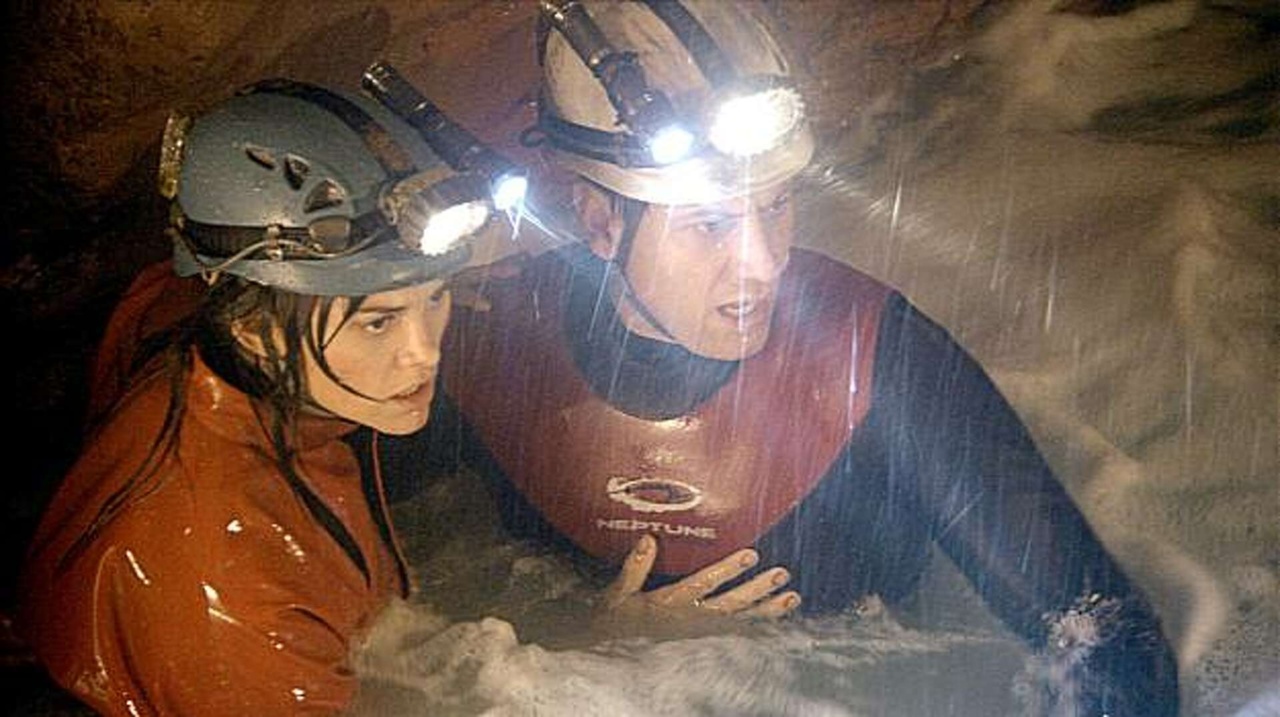 Two people wearing helmets and wetsuits, equipped with headlamps, navigate through a cave with rushing water around them, appearing cautious and focused.