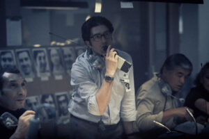 Three people in a dimly lit control room, one speaking into a handheld radio while others wear headsets, observe a wall with black-and-white photos in the background.