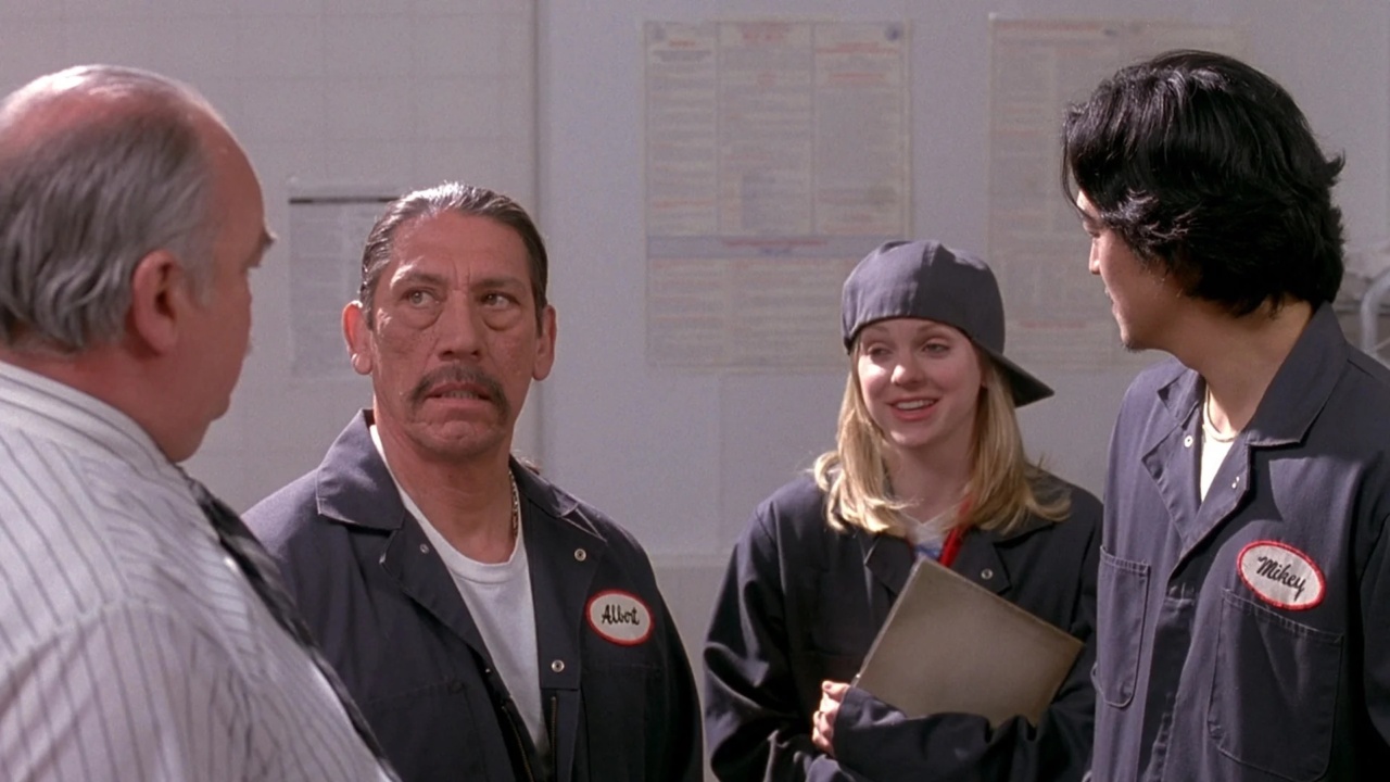 Four people wearing uniform shirts with name tags stand together, two facing away. One holds a folder and wears a backwards cap. They appear to be in a discussion in a workplace setting.