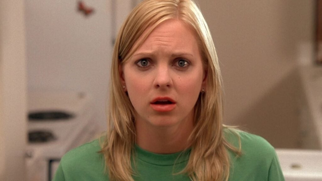 A person with long blonde hair, wearing a green top, looks surprised with slightly parted lips and raised eyebrows.