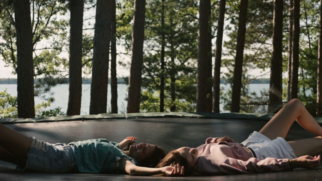 Two people lie on a trampoline in a forested area by a lake, wearing casual summer outfits.