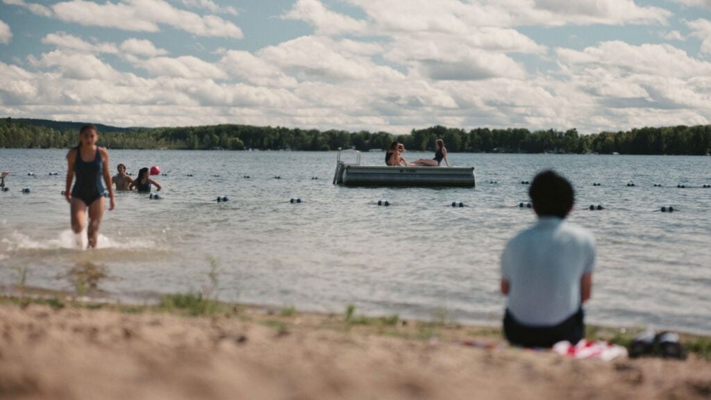 People enjoying a sunny day at a sandy lakeside beach, with some swimming and others sitting by the shore. A small dock floats on the water.