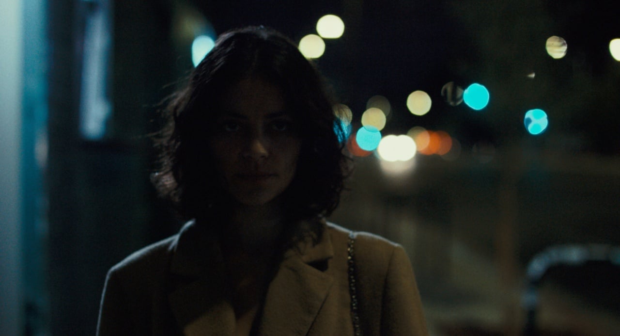 A person with curly hair stands outdoors at night, with blurred city lights in the background.