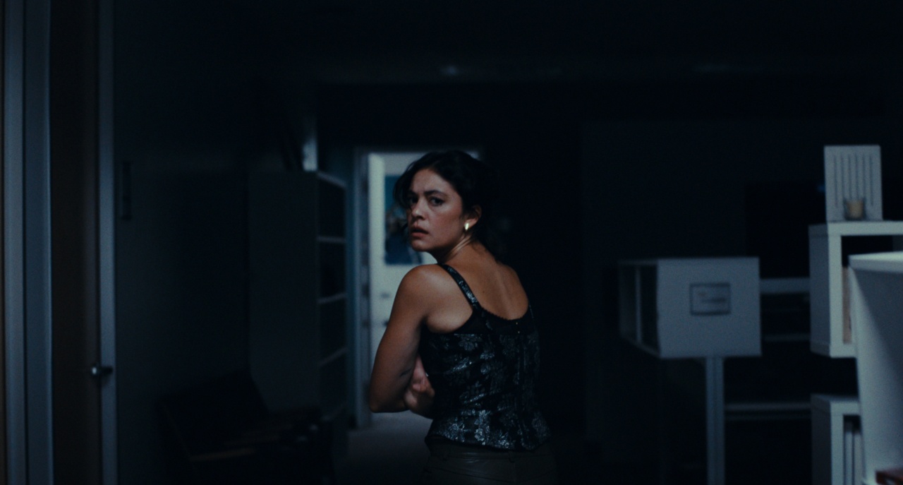 Woman in a dimly lit hallway looking over her shoulder, wearing a dark top and earrings.