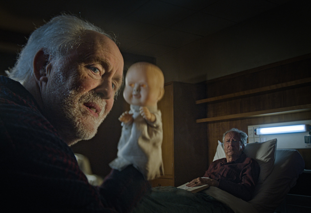 An elderly man holds a doll while another man lies in a hospital bed, staring towards the first man in a dimly lit room.