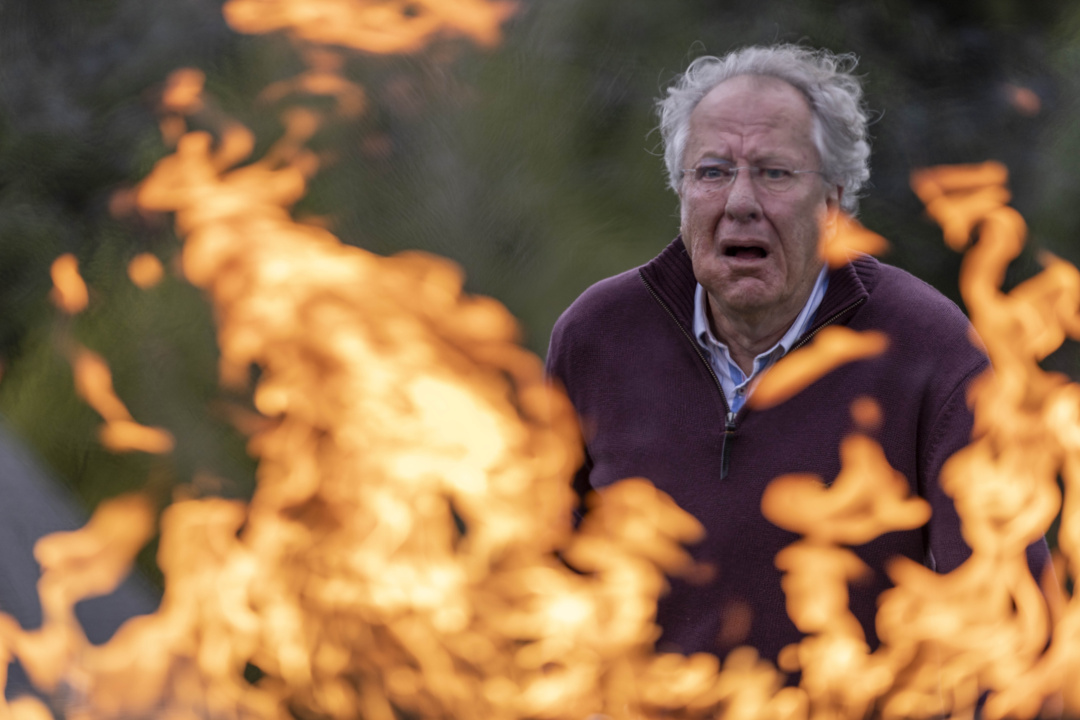 An elderly man stands behind large flames outdoors, looking distressed.