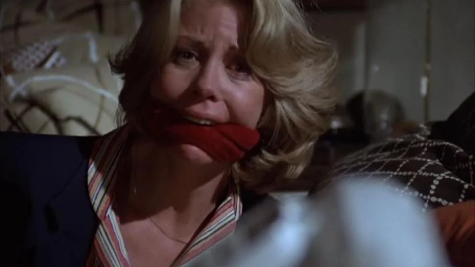 A woman with blonde hair is gagged with a red cloth in a dimly lit room, looking distressed.