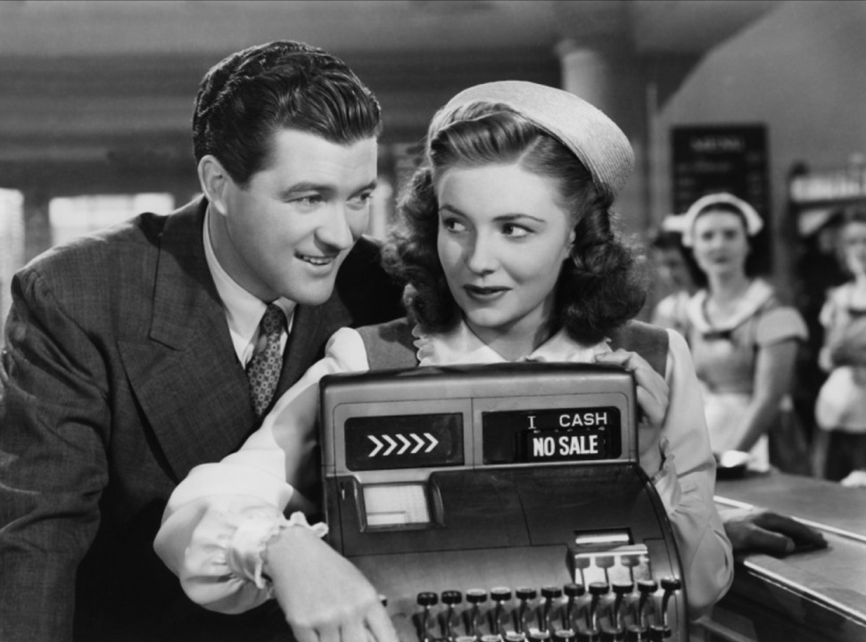 A man and woman stand at an old-fashioned cash register displaying "NO SALE." Two women are visible in the background, dressed in uniforms.