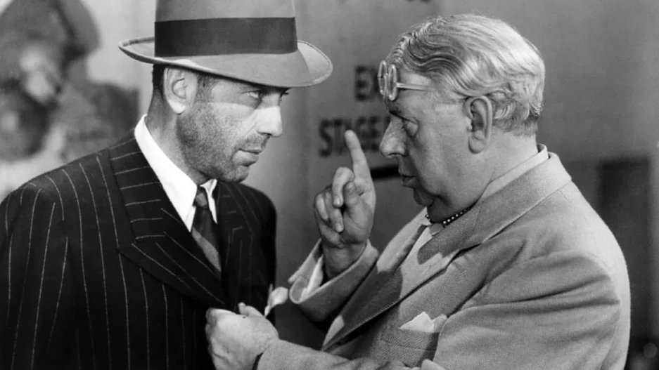 Two men in a black-and-white image. The man on the left wears a pinstripe suit and hat, while the man on the right, wearing glasses, points a finger at him.