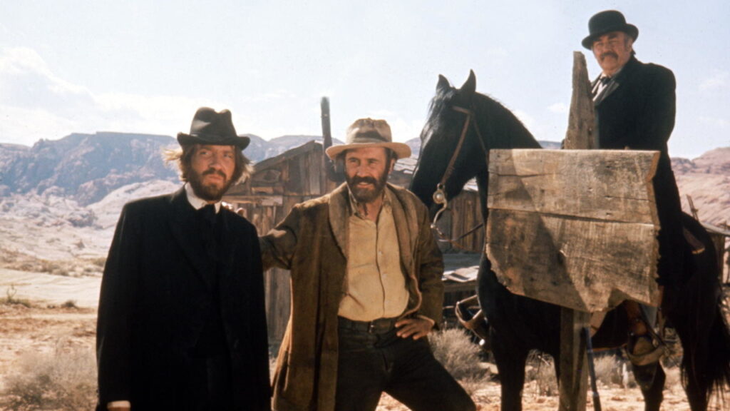 Three men in Western attire stand in a desert setting beside a wooden signpost and a horse, with a rustic building and rocky hills in the background.
