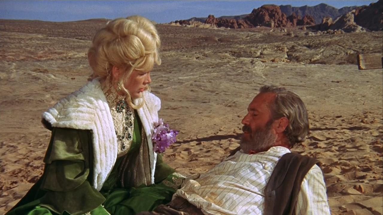 A woman in ornate green and white attire tends to a reclining man in a desert landscape.