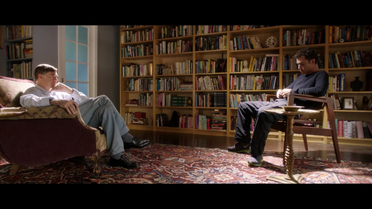 Two men sit in a room with a large bookshelf. One man is on a red sofa, the other on a wooden chair. A patterned rug covers the floor.