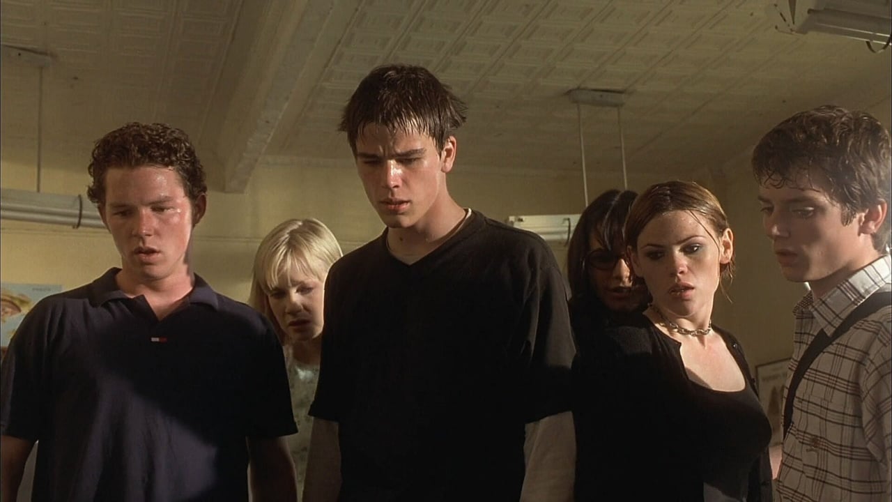A group of six young adults stand together, looking down with serious expressions in a dimly lit room.