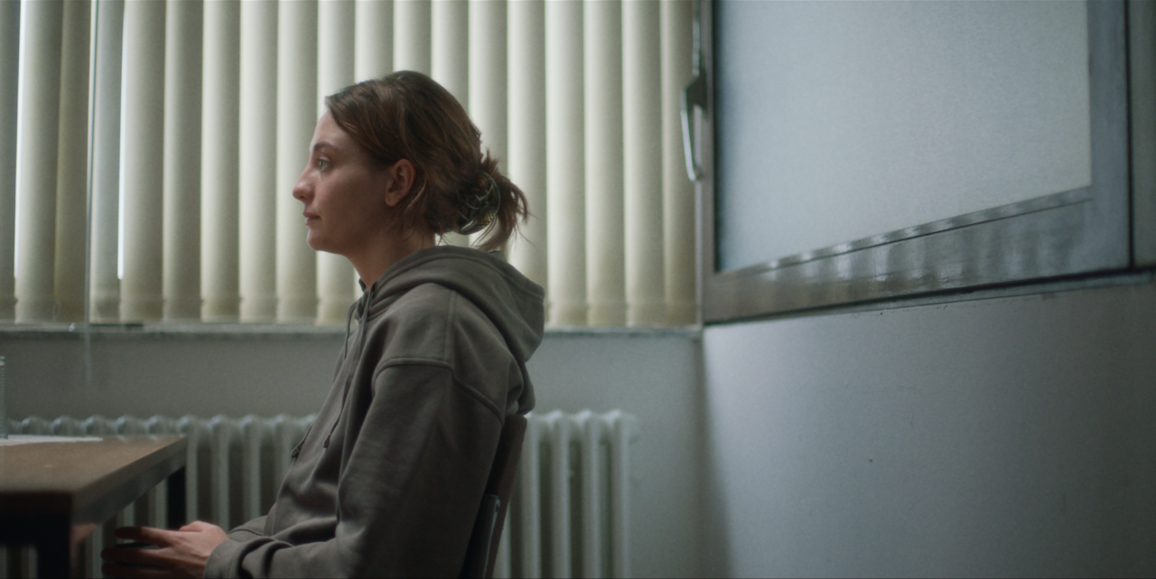 A person in a hoodie sits in profile in a room with vertical blinds and a radiator, looking forward.