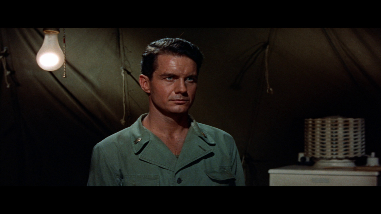 A man in military attire stands in a dimly lit room with a hanging light bulb and a ventilation device visible in the background.