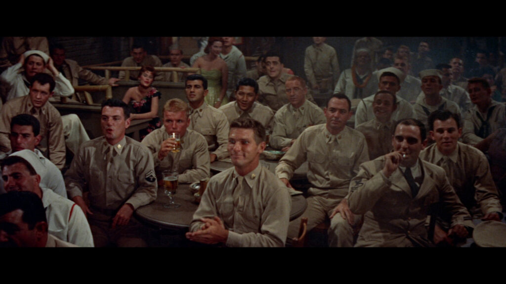A group of men in military uniforms sit at tables, watching a performance. One man holds a beer, and several have cigars. A woman in a red dress is visible in the background.