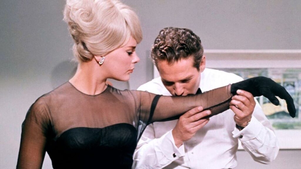 A woman in a sheer black dress sleeve stands while a man in a white shirt kisses her arm.