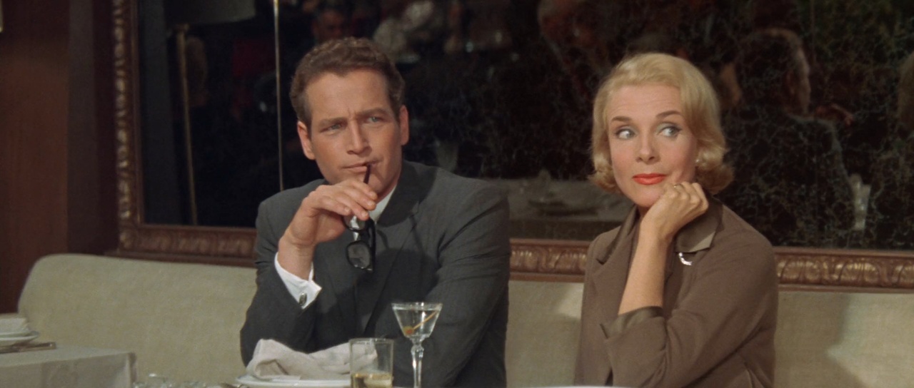 A man and woman sit together at a table in a restaurant. The man holds eyeglasses and a cigarette, and the woman rests her chin on her hand, looking sideways. There are drinks on the table.