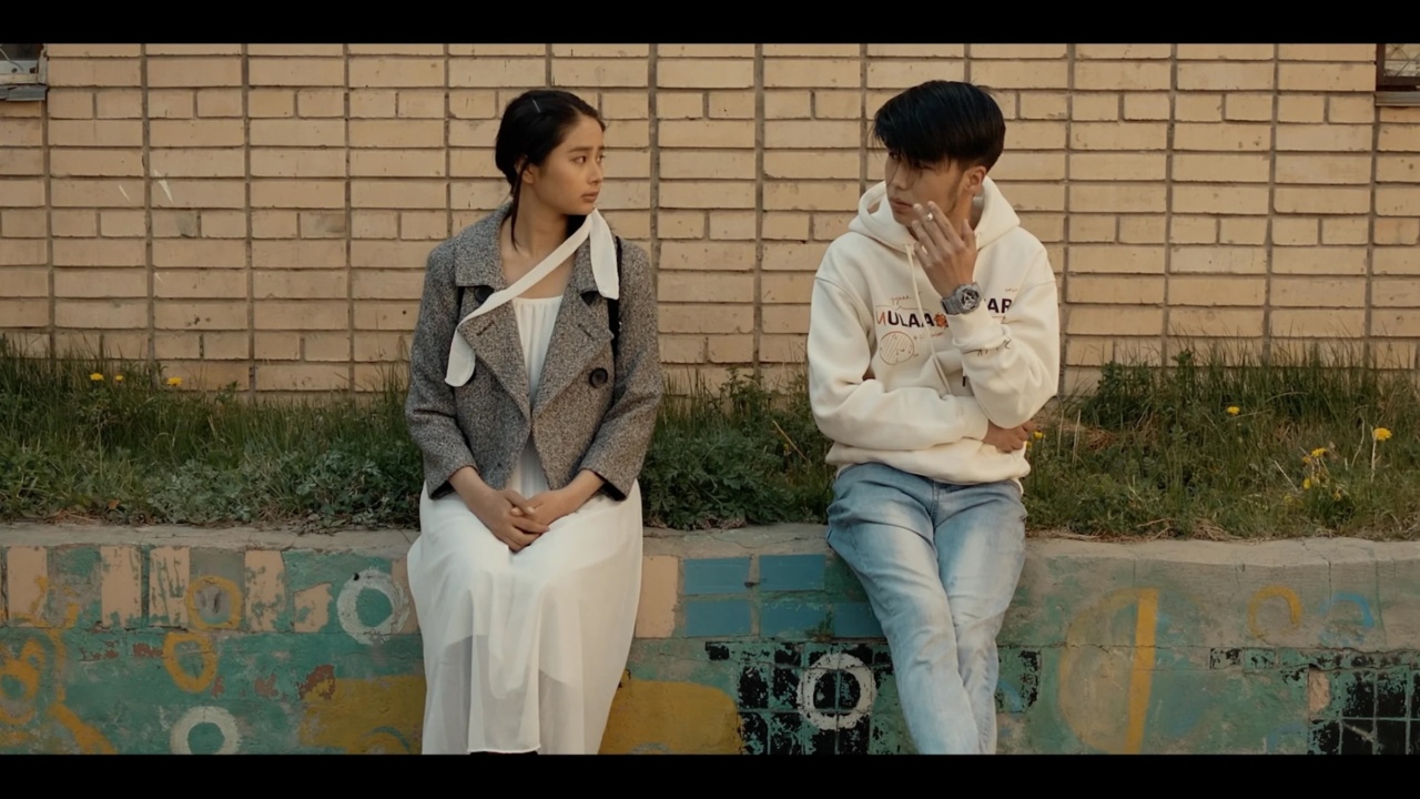 A woman in a coat and dress and a man in a hoodie and jeans sit on a graffiti-covered ledge against a brick wall, facing each other.