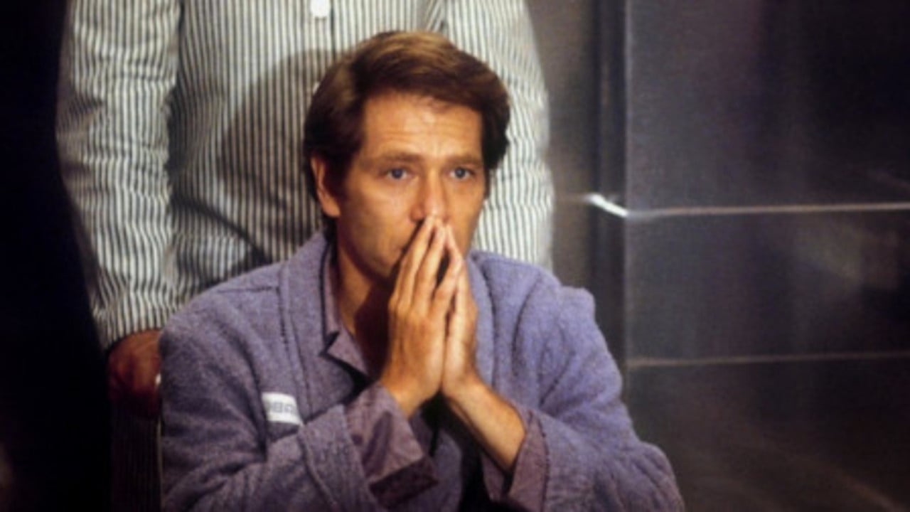 A person in a robe sits with hands clasped near their mouth, appearing contemplative or concerned. Another person stands behind, wearing striped attire.