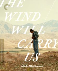 A person standing on a dirt path, looking down, with mountains in the background. Text on the image reads: "The Wind Will Carry Us, a film by Abbas Kiarostami.