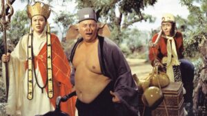 Three actors in elaborate costumes stand outdoors; one wears a crown and robe, another has a grey hat and exposed torso, and the third resembles a monkey with a red top.