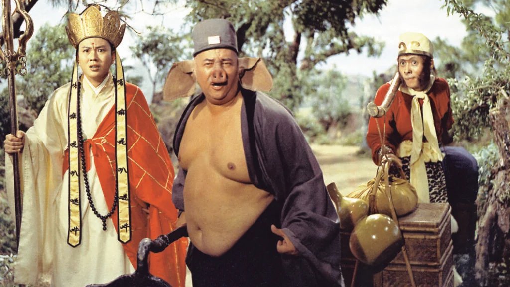 Three actors in elaborate costumes stand outdoors; one wears a crown and robe, another has a grey hat and exposed torso, and the third resembles a monkey with a red top.