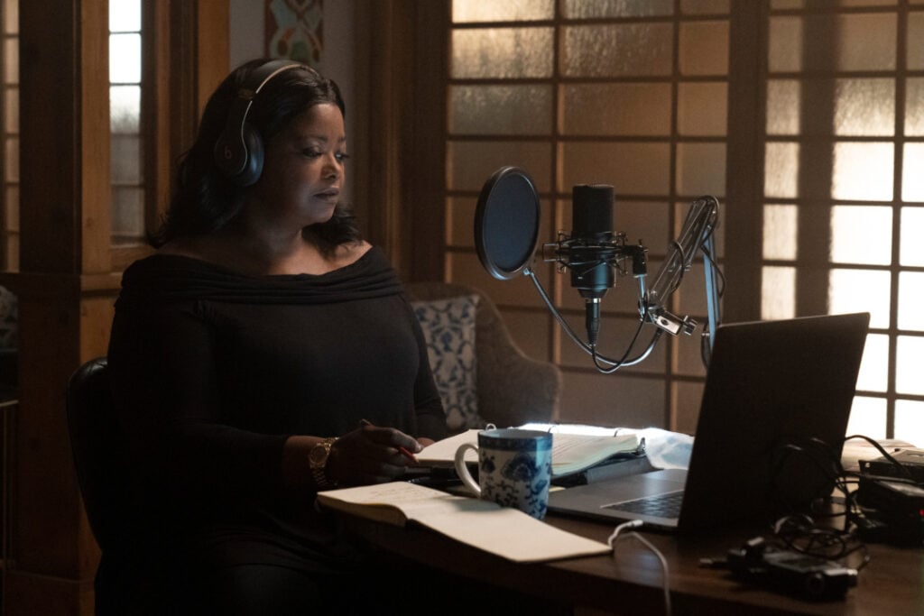 A person wearing headphones sits at a table with a microphone, laptop, and papers in front of them, appearing focused.