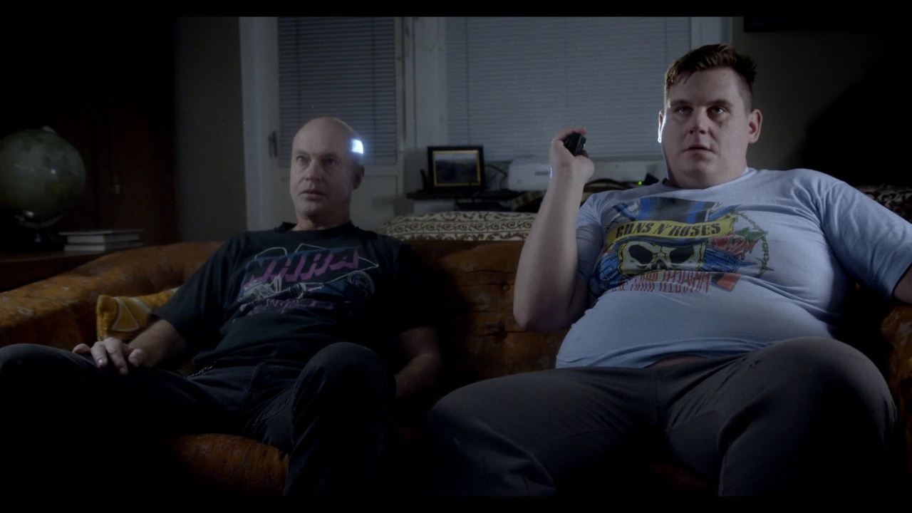 Two men sitting on a couch watching TV. The man on the left wears a black shirt, and the man on the right holds a remote, wearing a light blue shirt. They appear focused on the screen.
