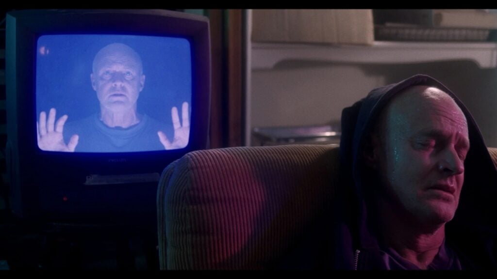 A man sitting in a chair with his eyes closed, while his image is shown on a TV screen beside him, with his hands raised.