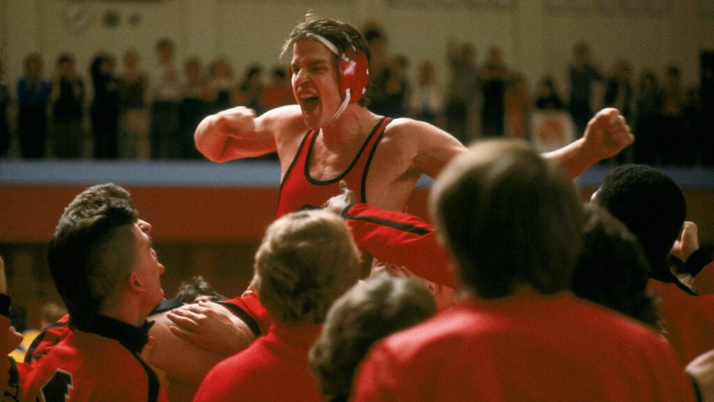 A wrestler in a red uniform and headgear is triumphantly raised by a crowd, celebrating a victory in a gymnasium. Spectators are visible in the background.