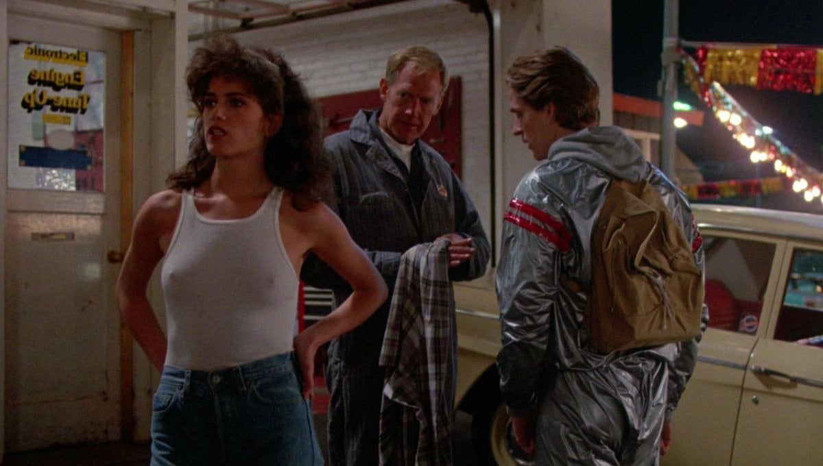 A woman in a white tank top stands in front of two men. One man holds a jacket, and the other wears a shiny silver suit with a backpack. A yellow car and festive banners are in the background.
