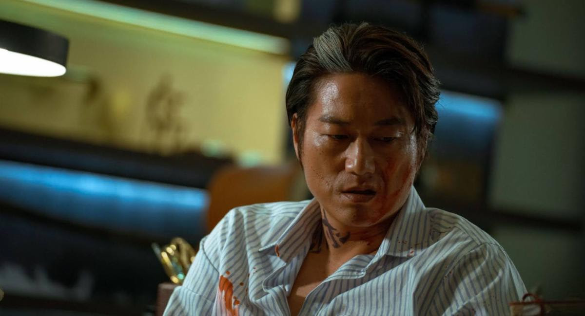 A person wearing a shirt with red stains looks downward in a dimly lit room.