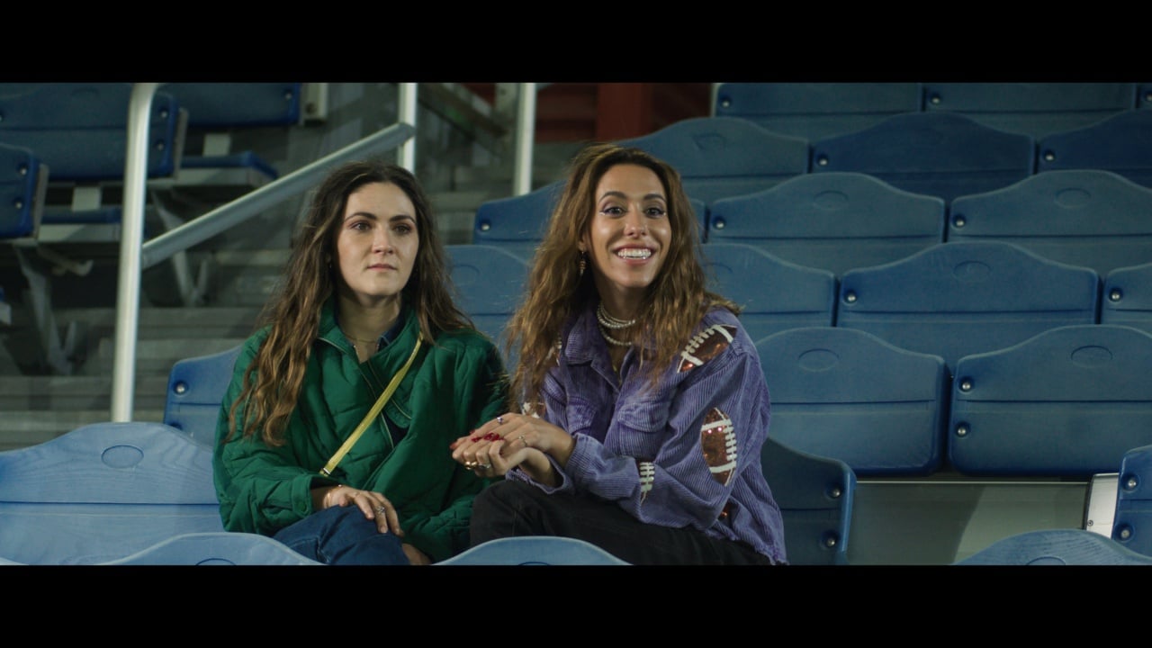 Two women sitting in blue stadium seats; one is smiling, and the other has a neutral expression.