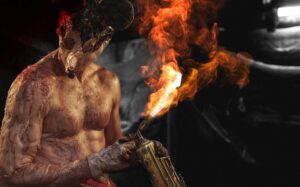 A shirtless person in a tattered costume uses a blowtorch, creating a flame, against a dark background.