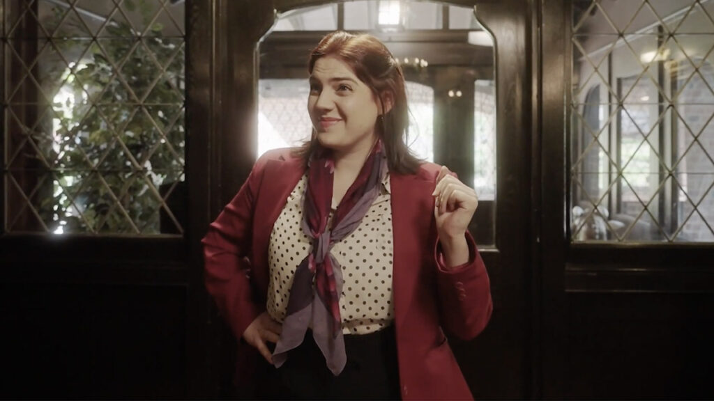 Woman in a red blazer and polka dot shirt stands in front of a windowed door, smiling with her right hand raised slightly.