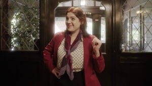 Woman in a red blazer and polka dot shirt stands in front of a windowed door, smiling with her right hand raised slightly.