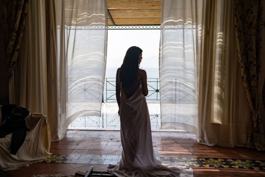A woman wrapped in a sheet stands in a room, facing a balcony with drawn curtains, overlooking a seaside view.