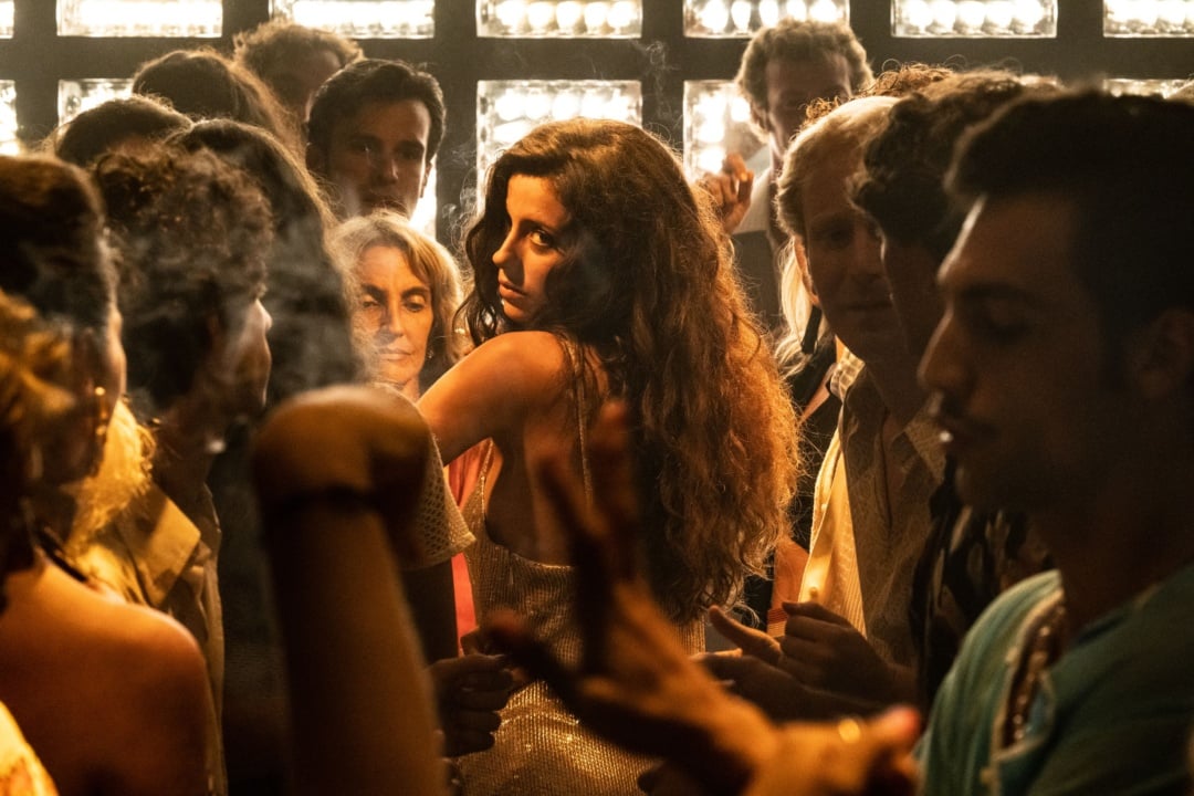 A woman in a glittery dress looks back amidst a crowded nightclub scene with people dancing and socializing under warm lighting.