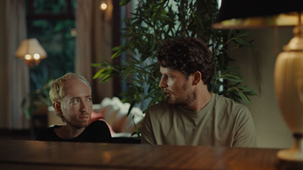 Two men sit next to each other indoors, engaging in conversation. A large plant and warm lighting decorate the background.