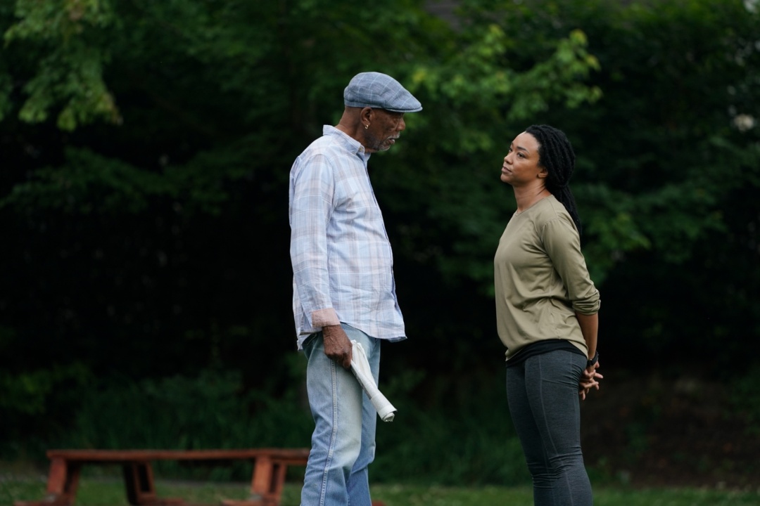 An older man and a younger woman stand facing each other outdoors. The man holds a rolled-up paper, and trees are visible in the background.