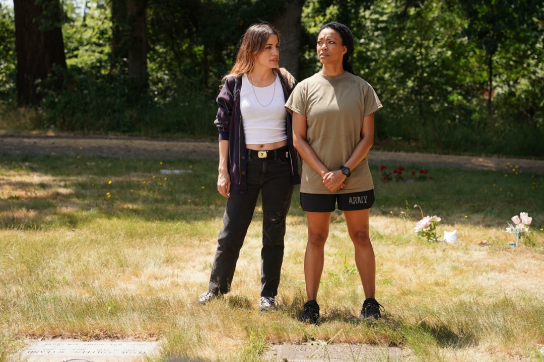 Two women stand on grass in a park; one in a plaid shirt and jeans, the other in an "Army" t-shirt and shorts. Trees and flowers are visible in the background.