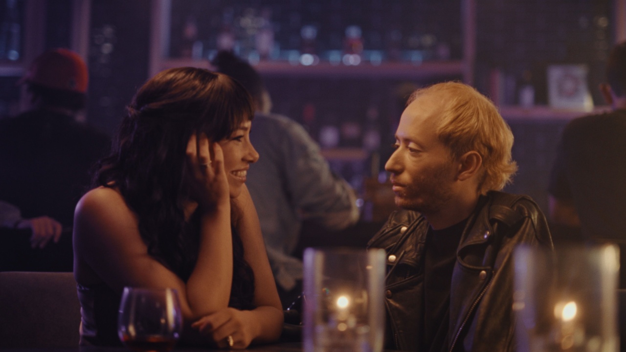 A woman and a man sitting at a dimly lit table in a bar, smiling at each other with drinks and candles on the table.