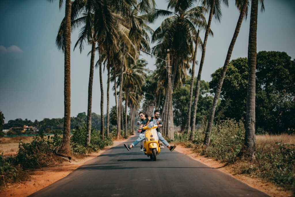 Two people ride a yellow scooter down a road lined with tall palm trees on a sunny day.