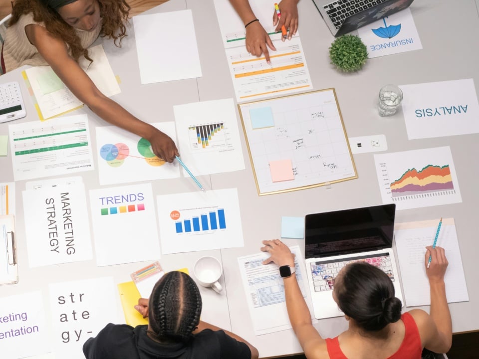 Top view of a meeting over a table with charts, graphs, and documents labeled marketing strategy, trends, analysis. Four people are discussing and pointing at papers and a laptop.