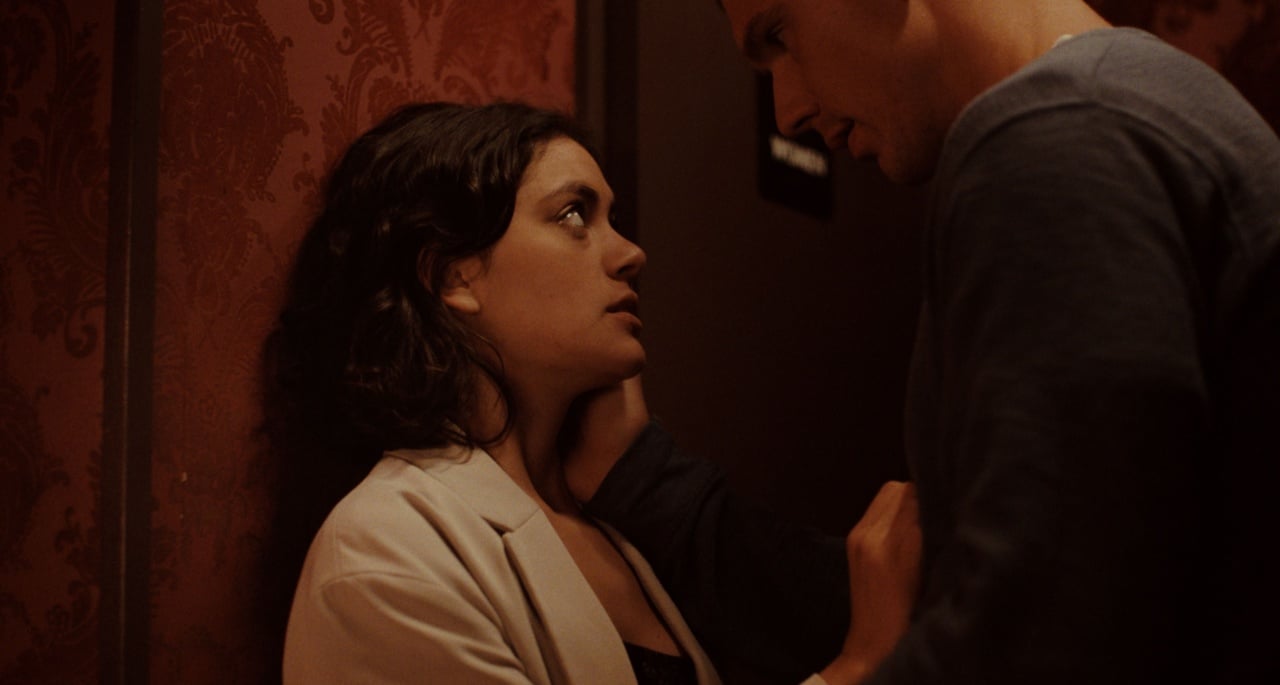 A woman leaning against a patterned wall looks up at a man who is gently touching her face. The setting is dimly lit and intimate.
