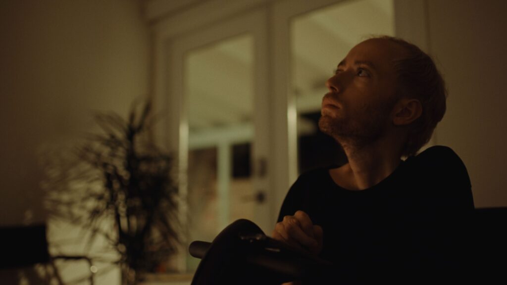 A person with short hair and a beard, wearing a dark shirt, sits indoors, looking upward. The room is dimly lit with a plant visible in the background near a window.